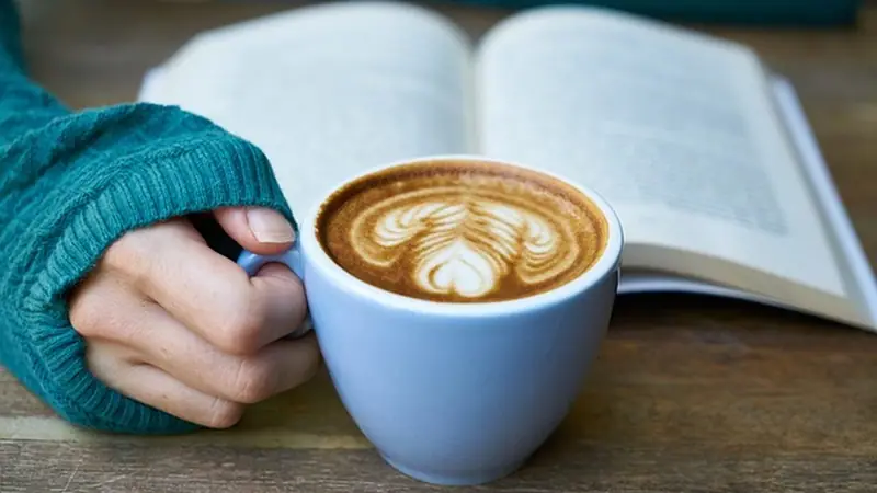 Manos con una taza de café junto a un libro abierto